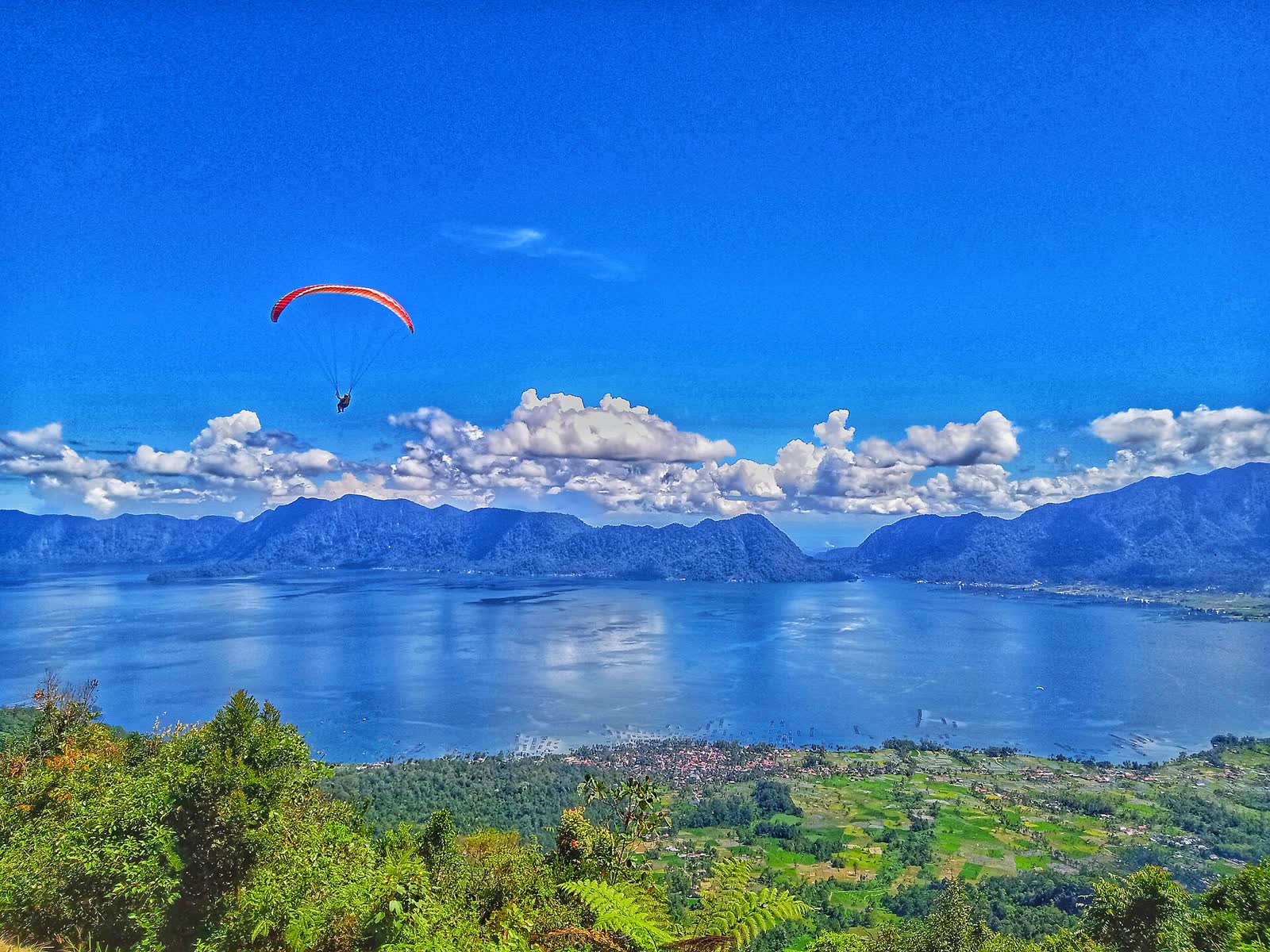 Paralayang merah di langit biru di atas Danau Maninjau, dilihat dari Lawang Top, Agam